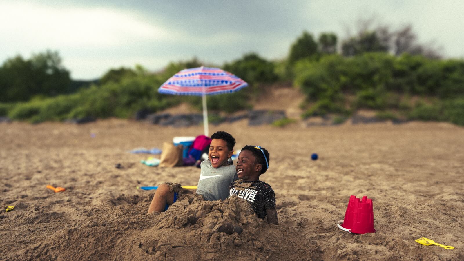 two young boys playin in the sand with a beach umbrella in the background
