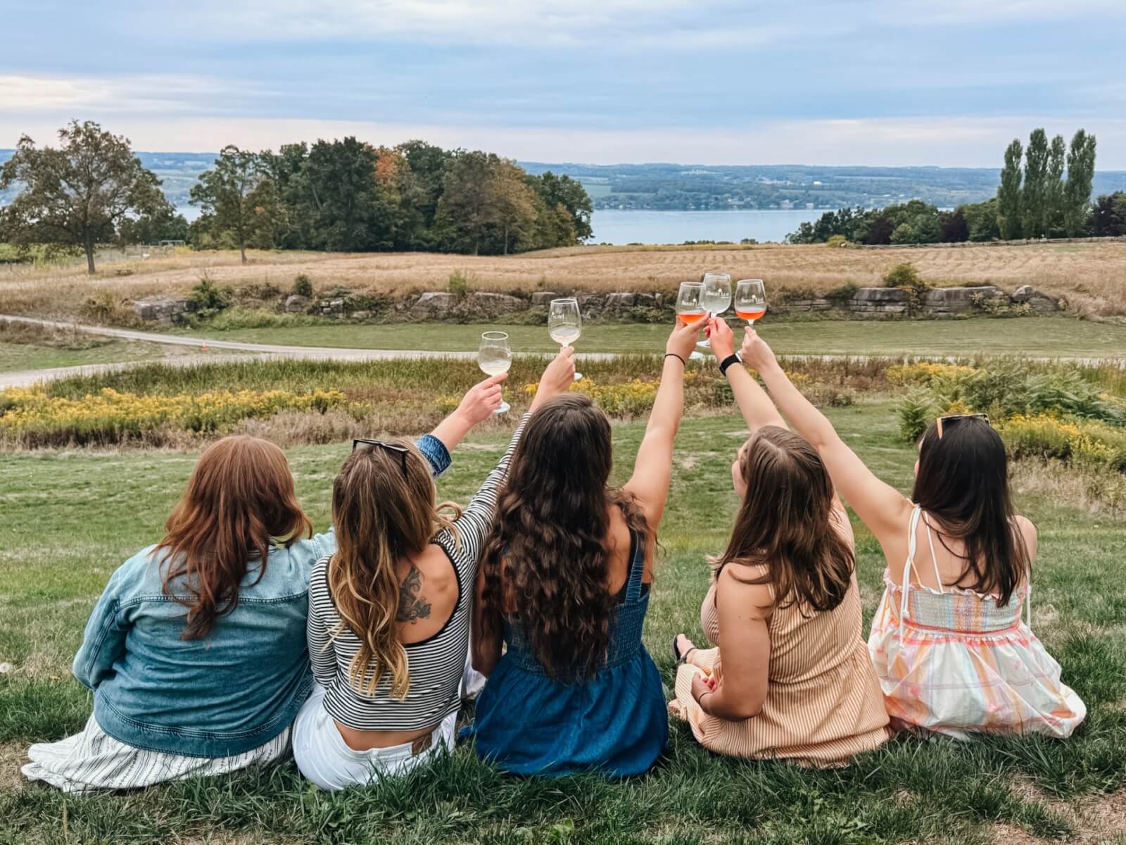 FIVE 
WOMEN SITTING ON THE GRASS, OVERLOOKING CAYUGA LAKE,  RAISING A GLASS OF WINE 