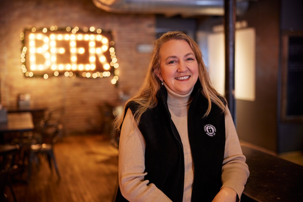 Women in Craft Beverage: Pioneers of Cayuga County owner of Prison City Brewery standing in front of a beer sign