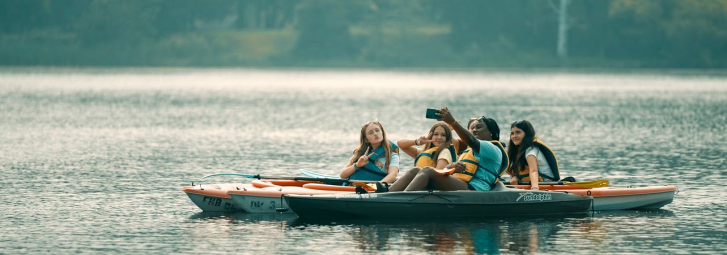 Five young girls sit on three kayaks on the water and take a selfie together.