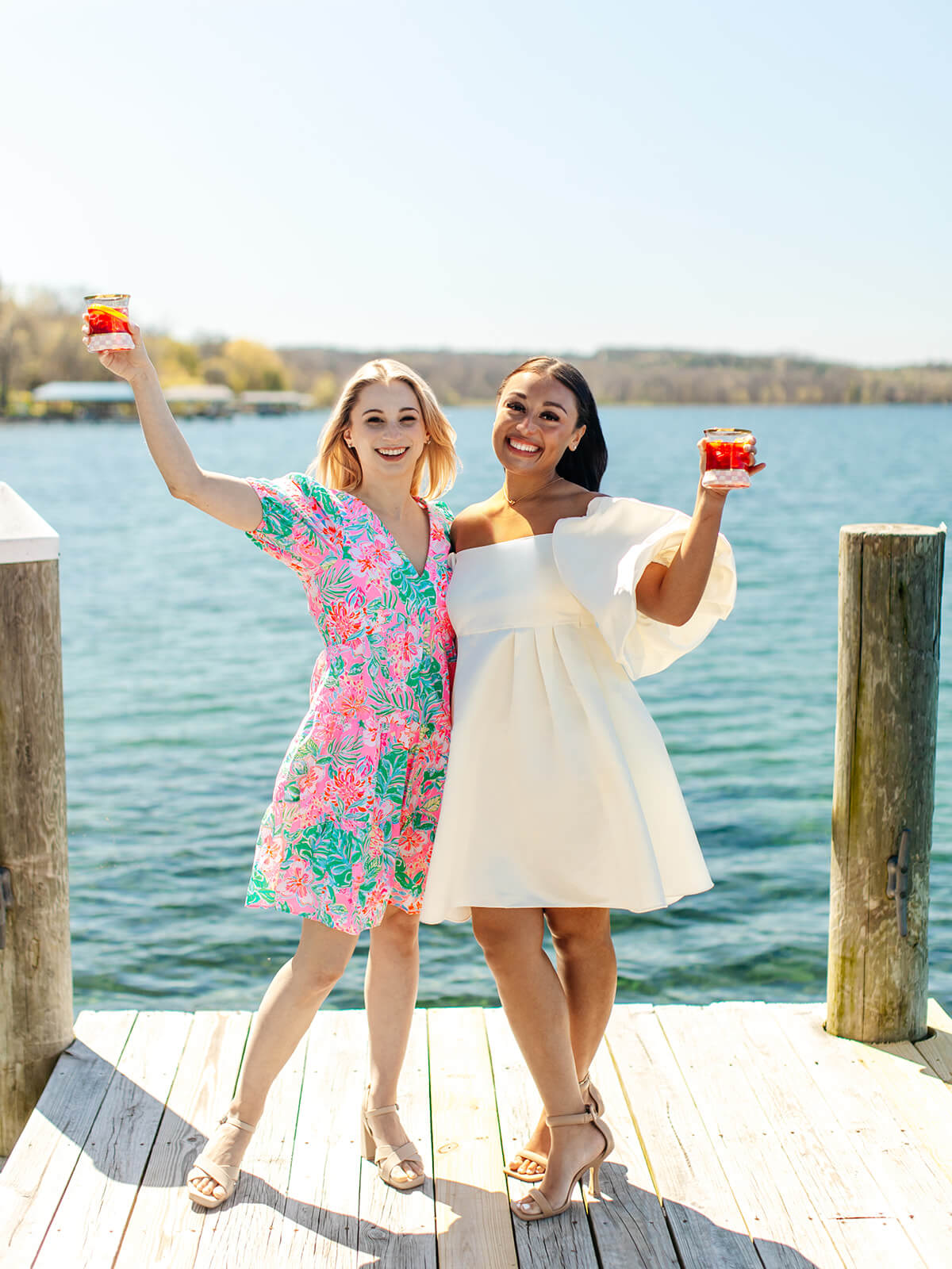 two women standing on a dock with Cayuga Lake in the background holding a cocktail and toasting each other