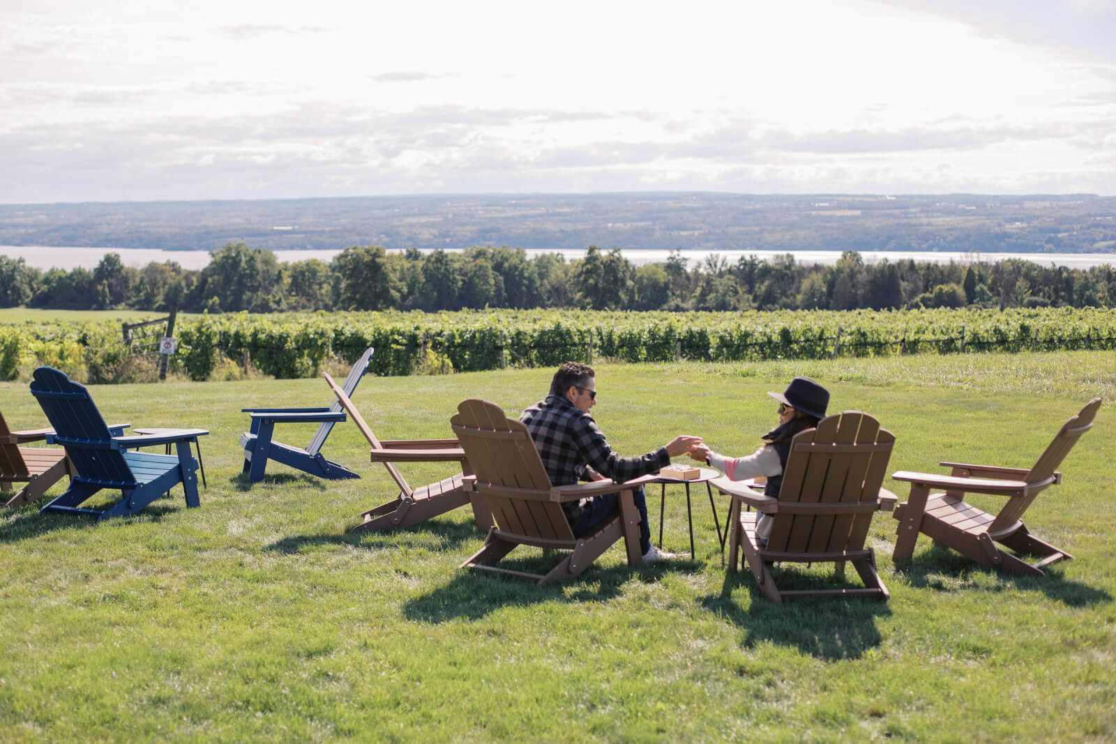 Who benefits when visitors come to Cayuga County? visitors to a winery sitting in lounge chairs enjoying a glass of local wine and the scenery of a lake and grape vines
