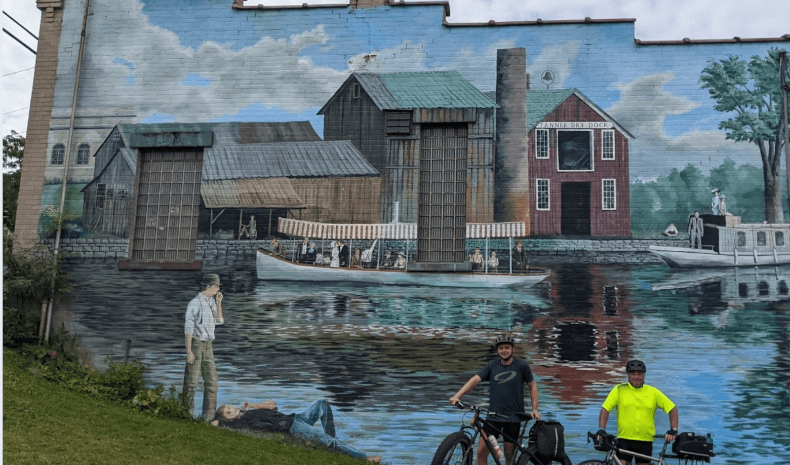 Image of a mural of the Erie Canal with cyclists in front. Mural located in the town of Port Byron